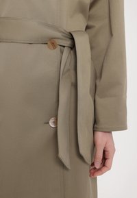 Gros plan d'une personne portant un manteau beige avec une ceinture nouée à la taille et deux boutons marron visibles.