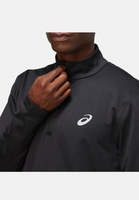 Black long-sleeve pullover with a high collar, featuring a half-zip. Reflective logo on the chest; smooth, stretchy fabric.