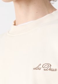 Gros plan d'un sweat-shirt à col rond couleur crème avec « Les Deux » brodé en écriture cursive marron sur la poitrine.