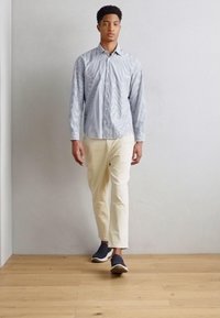 En knappmessig skjorte med blå og hvite vertikale striper, lyse beige bukser og mørke slip-on sko. Står foran en enkel grå vegg og tregulv.