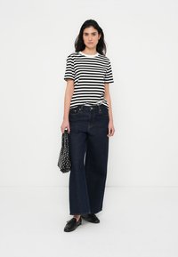 Black and white striped t-shirt, high-waisted dark denim wide-leg pants, black shoes, holding a polka dot bag. Minimalist design.