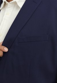 Marinblazer aus texturiertem Stoff, mit einer Brusttasche und Reverskragen, darunter ein weißes Hemd mit Knöpfen.