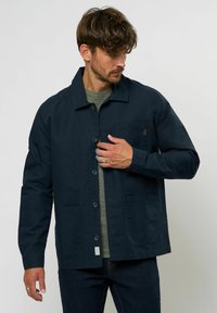 Chaqueta azul marino de algodón, con cierre de botones, dos bolsillos frontales y un pequeño logo en el bolsillo del pecho. Capa interior texturizada.