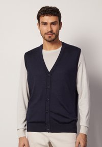 Mörkblå stickad väst med v-ringning och sex knappar, som bärs över en ljusfärgad långärmad skjorta. Slät textur och figurnära design.