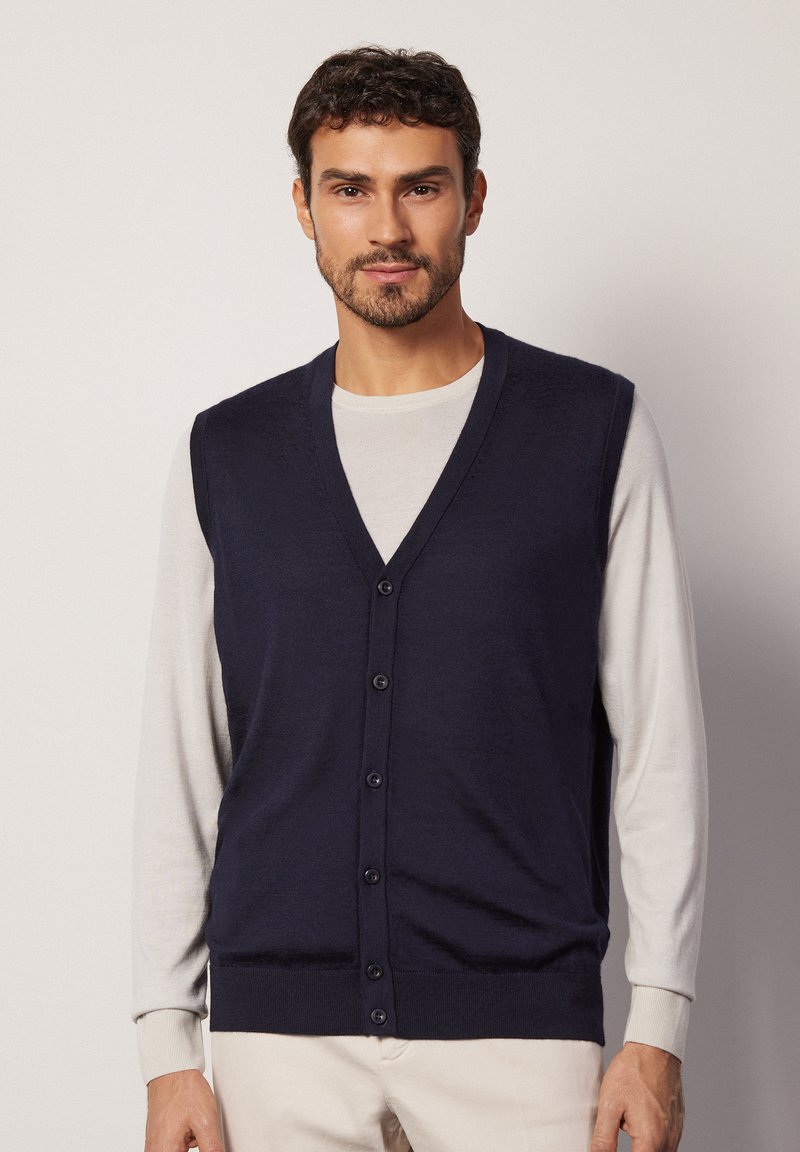 Mörkblå stickad väst med v-ringning och sex knappar, som bärs över en ljusfärgad långärmad skjorta. Slät textur och figurnära design.