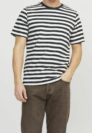 Uomo che indossa una maglietta a maniche corte a strisce orizzontali bianche e nere e pantaloni marroni, con una mano in tasca.