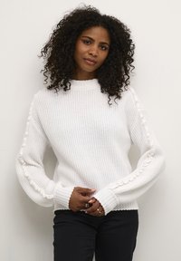 Maglione bianco lavorato a maglia con scollo rotondo, polsini a costine e dettagli testurizzati lungo le maniche. Indossato con pantaloni neri, mani incrociate.