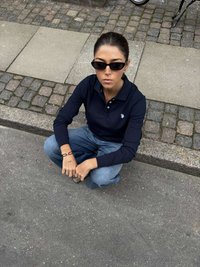 Tricou polo cu mâneci lungi de culoare bleumarin, cu emblemă, asortat cu blugi deschis la culoare. Persoană așezată, purtând ochelari de soare negri, în fața unui fundal de piatră cubică.