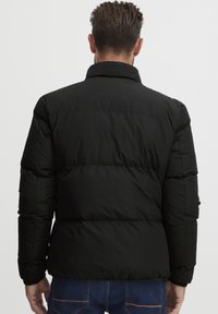 Črna napihljiva jakna z visokim ovratnikom in šivanim panelom. Ima elastične manšete in eleganten, prešit teksturiran videz. Idealna za hladno vreme.