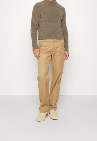 Bruine gebreide trui, tan kleurige broek met zichtbare stikdetails, en beige enkellaarsjes. Soepele texturen van de stof overal.