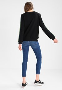 Czarna bluza z tęczowymi paskami na rękawach, zestawiona z niebieskimi wąskimi dżinsami i czarnymi sneakersami, widziana z tyłu.