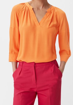 Vrouw draagt een feloranje V-hals blouse met driekwart mouwen, ingestopt in hooggetailleerde, op maat gemaakte roze broek met handen in de zakken.