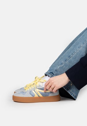 Person in blauen Jeans passt hellblaue und gelbe Sneakers mit Gummisohlen vor weißem Hintergrund an.