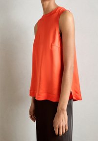 Top rosso senza maniche realizzato in tessuto liscio, con scollatura rotonda, orlo leggermente svasato e tasca laterale. Abbinato a pantaloni scuri.