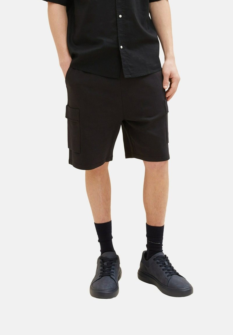 Osoba v černých cargo šortkách, černých ponožkách a černých teniskách, stojící s rukama v kapsách na bílém pozadí.