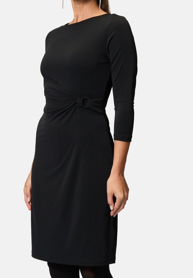 Femme portant une robe noire ajustée avec des manches trois-quarts et un détail de nœud torsadé à la taille, debout devant un fond uni.