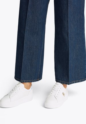 Weiße Sneaker mit glattem Lederfinish, flacher Laufsohle und auffälliger Schnürung, kombiniert mit dunkelblauen Jeanshosen mit weitem Bein.