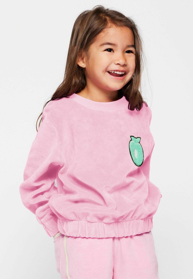 Sweatshirt en velours rose avec un patch rond de fruit vert sur la poitrine. Doté de poignets et d'un bas élastiqués pour un ajustement confortable. Texture douce.