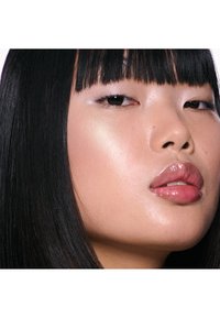 Close-up van een gezicht met een gladde, glanzende huid, volle lippen en steil zwart haar met rechte pony. Subtiele highlighter op de wangen en oogleden.