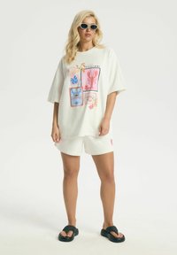Kobieta w oversize białej koszulce z kolorowymi nadrukami owoców morza, białych szortach, czarnych sandałach i okularach przeciwsłonecznych stojąca na białym tle.