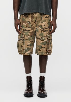 Personne portant un short cargo camouflage vert et marron, des bottes à lacets marron foncé et des chaussettes blanches, debout avec les mains détendues le long du corps.