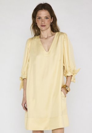 Femme aux cheveux châtain clair portant une robe ample jaune pâle à col en V et manches 3/4 nouées, accessoirisée avec des bracelets et des boucles d'oreilles créoles.
