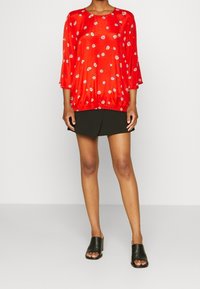 Blusa roja de flores con mangas de tres cuartos, dobladillo fruncido, textura brillante, llevada sobre una mini falda negra. Unos zapatos deslizantes negros completan el look.