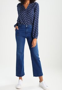 Chemisier bleu marine avec motif ovale blanc, col en V et poignets boutonnés, associé à un jean en denim taille haute à jambe large et des baskets blanches.
