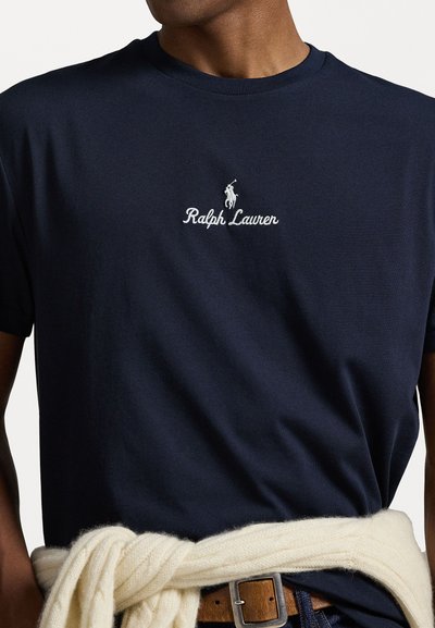 T-shirt de algodão azul marinho com mangas curtas, apresentando um logótipo branco bordado e o texto "Ralph Lauren" no peito. Sobreposto com um suéter de malha em creme amarrado na cintura.