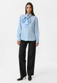 Lichtblauwe blouse met een grote strikdetail bij de kraag, knopen aan de voorkant en lange mouwen. Gecombineerd met donkere wijde jeans en zwarte hakken.