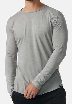 Homme portant un pull gris chiné à manches longues avec un pantalon noir, debout, une main près de la taille et l'autre dans la poche.