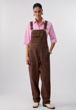 Donna con salopette marrone e maglietta a righe rosa, in piedi con una mano in tasca, stivali bianchi e braccialetti d'argento.