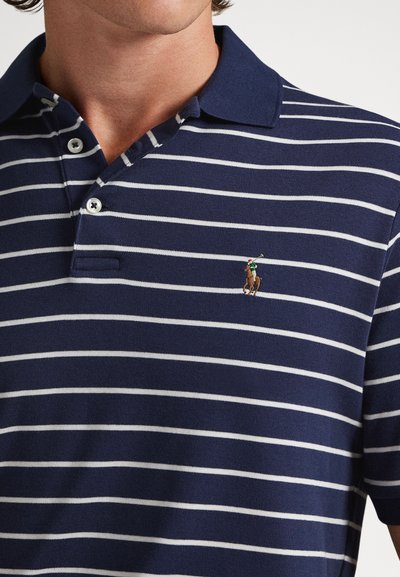 Vīrietis, kurš valkā tumši zilu un baltu horizontāli svītrainu polo krekliņu ar divām pogām un mazu izšūtu polo spēlētāja logo uz krūtīm.