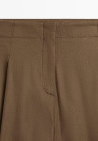 Close-up van de tailleband van een bruine broek en het bovenste voorstuk met zichtbare stiksels en ritssluiting.