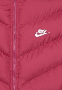 Bordo pūkų striukė su chevrono siuvinėjimu, priekinė užtrauktukas ir baltas Nike logotipas. Lygus paviršius ir lengvas audinys.