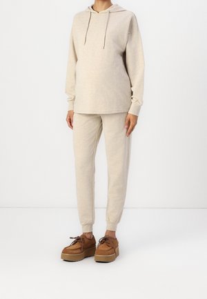 Osoba ubrana w beżową bluzę z kapturem i pasujące spodnie dresowe, stojąca na tle białego tła, w brązowych platformowych butach na sznurowadła.