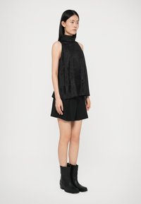 Zwart mouwloos topje met een hoge hals, losse fit en textuurstof, gecombineerd met zwarte shorts en zwarte enkellaarsjes.