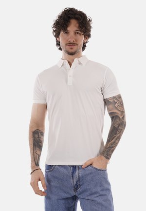 Uomo con capelli ricci che indossa una polo bianca e jeans blu, mostra tatuaggi su entrambe le braccia, in piedi davanti a uno sfondo semplice.