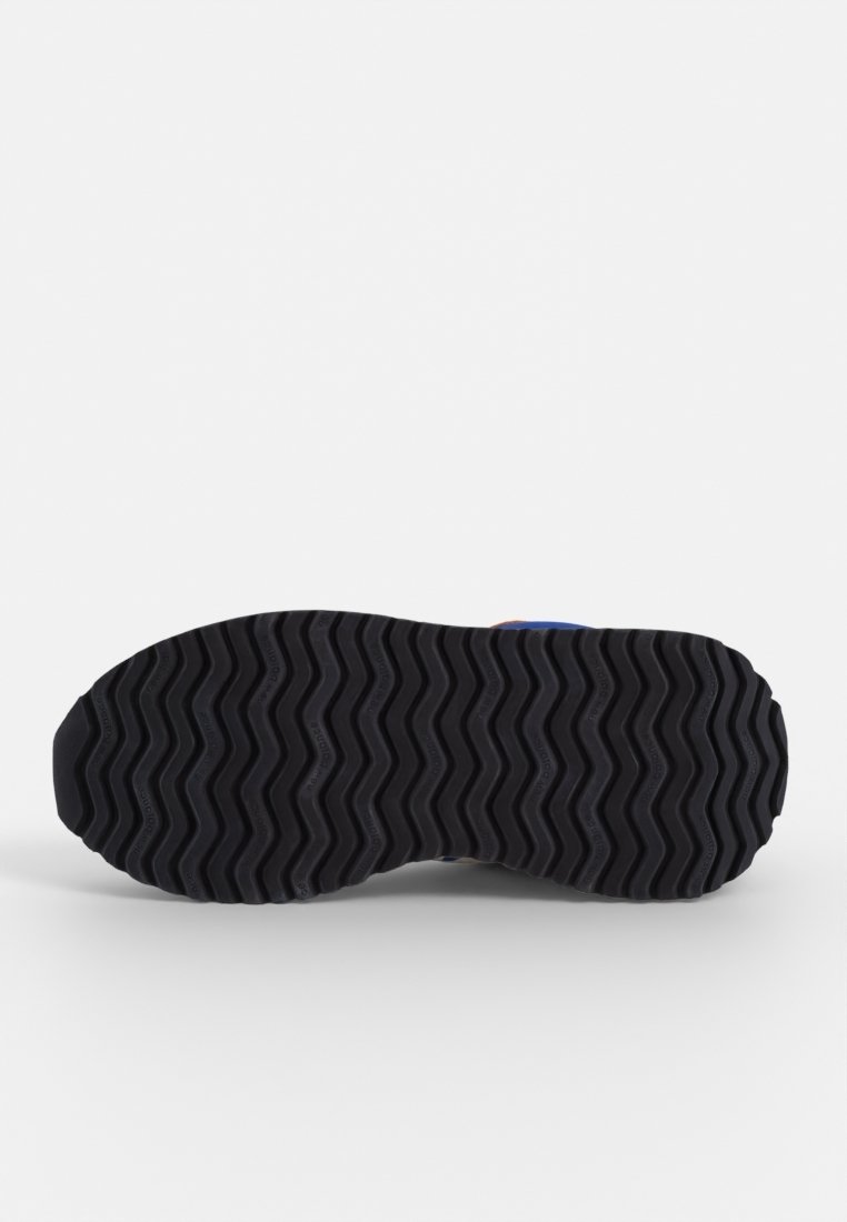 Semelle en caoutchouc noir d'une basket avec un motif de bande de roulement en zigzag, montrée à plat sur un fond blanc.