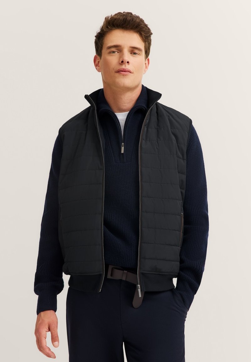 Schwarze gesteppte Weste mit hohem Kragen, Reißverschluss vorne und seitlichen Taschen, über einem dunkelblauen Pullover getragen. Glatte, leichte Textur.
