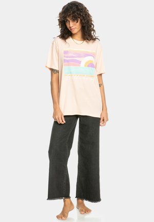 T-shirt oversize rosa chiaro con una grafica che presenta onde pastello e un sole, abbinata a jeans neri a gamba larga a vita alta.