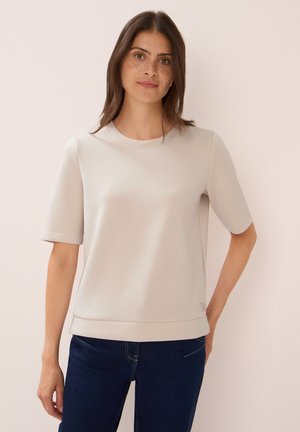 Frau mit langen braunen Haaren und Sommersprossen, die ein beiges Kurzarmoberteil und dunkelblaue Jeans trägt, steht vor einem hellen Hintergrund.