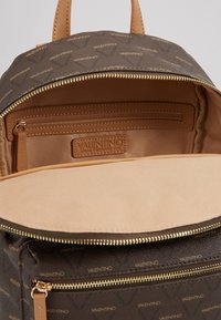 Mochila de couro castanho com textura suave, zíperes dourados e um padrão discreto de logótipos por toda a parte. O interior inclui um bolso com zíper e um bolso de deslizamento.