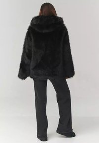 Czarna kurtka z sztucznego futra z kapturem o pudełkowym kształcie i miękkiej teksturze, zestawiona z czarnymi spodniami typu wide-leg i czarnym obuwiem.