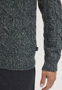 Grüner gestrickter Pullover mit einem strukturierten Zopfmuster. Hat gerippte Bündchen und Saum und zeigt ein Etikett an der Seite.