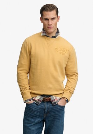Mann mit zurückgekämmtem Haar trägt einen senffarbenen Pullover über einem karierten Hemd, das in blaue Jeans gesteckt ist, Hände in den Taschen, neutraler Hintergrund.