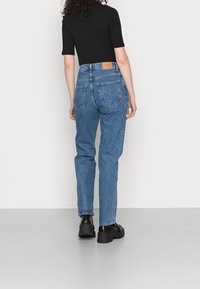 Jeans de denim azul de cintura alta con un corte recto, cinco bolsillos y una etiqueta de cuero marrón en la parte trasera de la cintura. Zapatos negros completan el look.