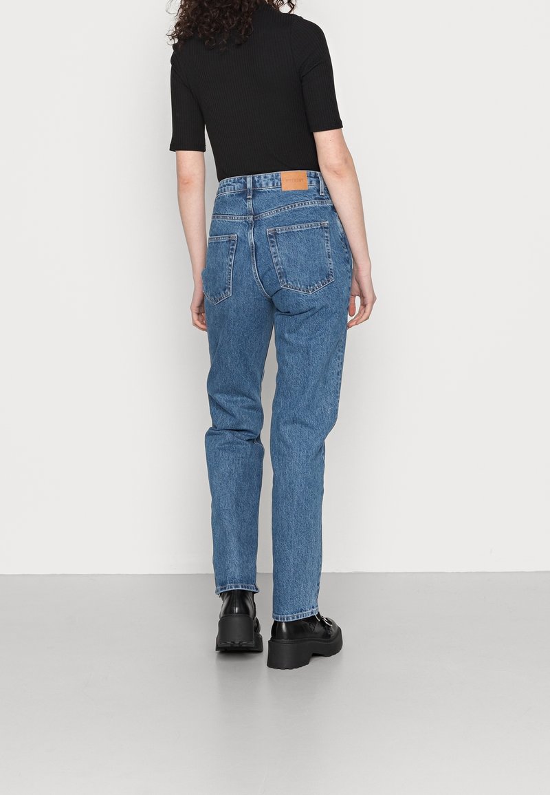 Jeans de denim azul de cintura alta con un corte recto, cinco bolsillos y una etiqueta de cuero marrón en la parte trasera de la cintura. Zapatos negros completan el look.