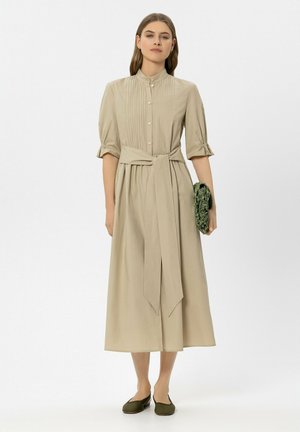 Frau trägt ein beiges Midikleid mit Knöpfen vorne, gebundenem Taillengürtel, gepufften Ellbogenlangen Ärmeln, hält eine strukturierte grüne Clutch und trägt grüne Ballerinas.