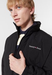 Giacca nera trapuntata con chiusura a zip, polsini elasticizzati e logo "Tommy Jeans". Indossata sopra una camicia a righe con collo alto.